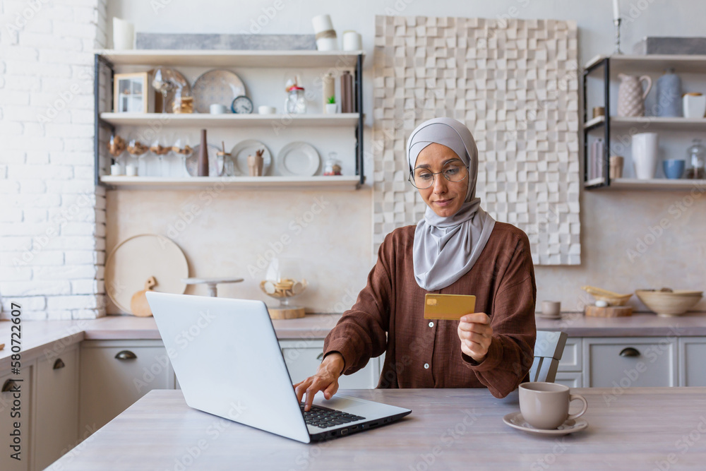 Young Muslim woman in hijab working remotely online. Sitting in the kitchen typing on a laptop and using a credit card. Online shopping, payment, transactions, investments, accounts. Young Muslim woman in hijab working remotely online. Sitting in the kitchen typing on a laptop and using a credit card. Online shopping, payment, transactions, investments, accounts.