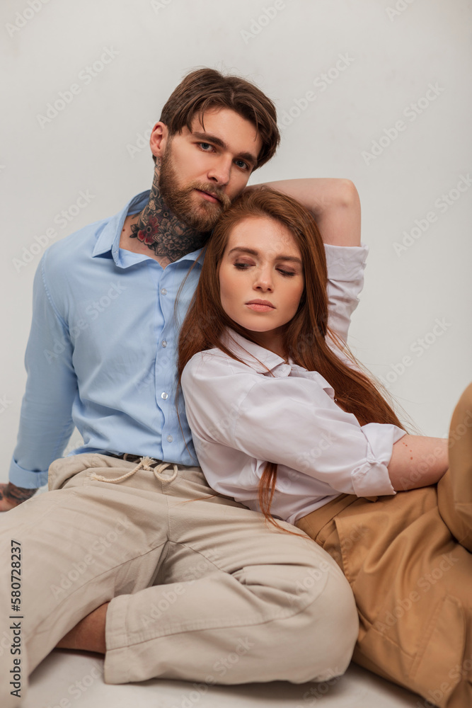 Fashionable beautiful caucasian couple in trendy clothes sits in the studio and poses on a white background. Beautiful young redhead girl and handsome hipster man Fashionable beautiful caucasian couple in trendy clothes sits in the studio and poses on a white background. Beautiful young redhead girl and handsome hipster man