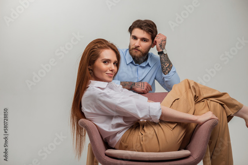 Fashion couple of lovers. Beautiful happy redhead girl model with red hair sits and rest in a chair and handsome brutal hipster man with beard and tattoo