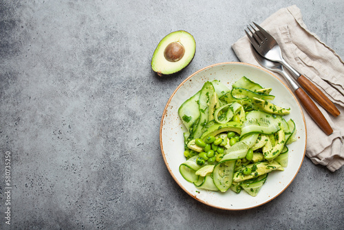 Healthy vegan green avocado salad bowl with sliced cucumbers, edamame beans, olive oil and herbs on ceramic plate top view on grey stone rustic table background. Space for text