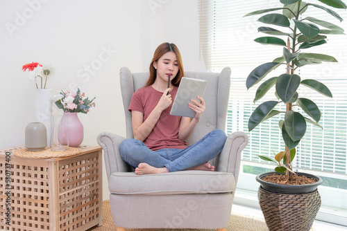 Asian woman sitting on yellow sofa using digital tablet and pen for learning shopping or working online near window