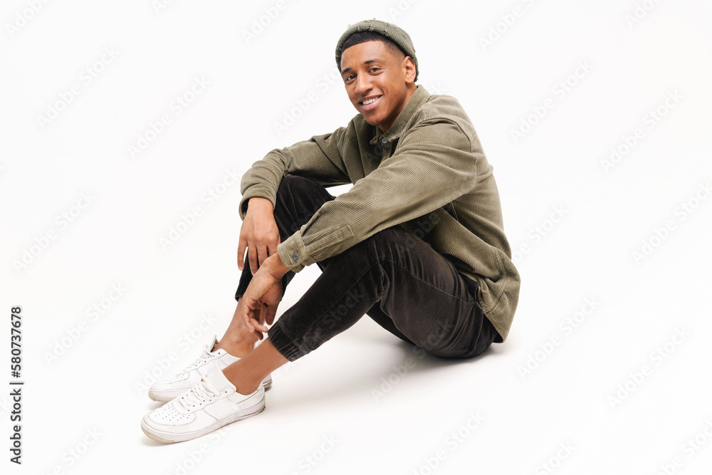 Naklejka premium Full length photo of carefree smiling african-american guy in hat sitting on the floor in relaxed pose isolated on white background