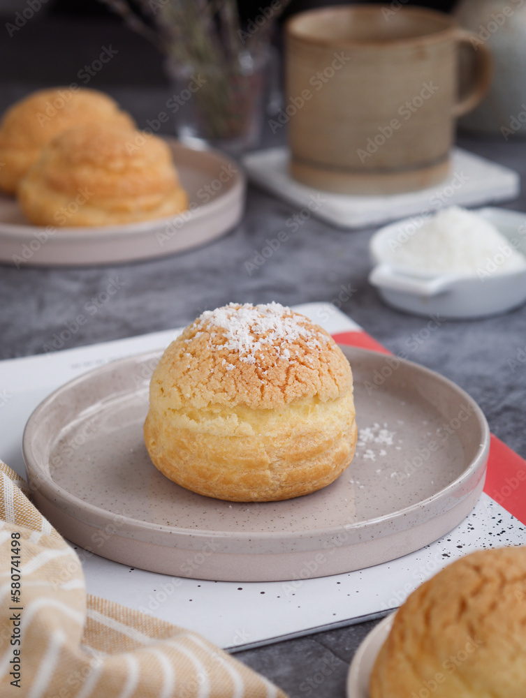 Close up of French cream puff crack bun (Choux au Craquelin) in a plate, topped with grated coconut filling with cup of coffee in background. It's a dessert base of savory choux dough bun Close up of French cream puff crack bun (Choux au Craquelin) in a plate, topped with grated coconut filling with cup of coffee in background. It's a dessert base of savory choux dough bun