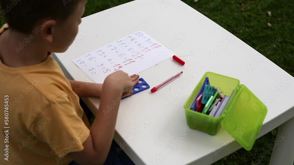 Child with autism has math therapy lesson: uses colorful Numicon shapes, counts, writes answers on paper; sits at white table in sunny garden with bright green grass Child with autism has math therapy lesson: uses colorful Numicon shapes, counts, writes answers on paper; sits at white table in sunny garden with bright green grass