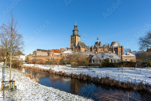 Canal and medieval wall of Hanseatic Dutch tower town Zutphen in The Netherlands covered in snow with historic heritage buildings against a clear blue sky. Winter wonderland picturesque scene.