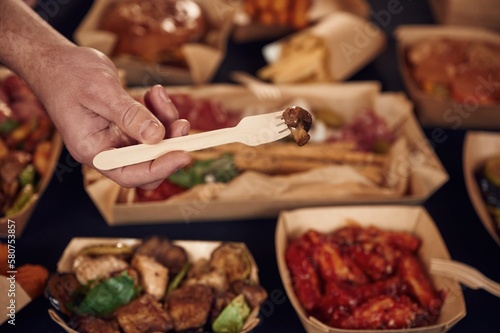 Man is holding fork with mushroom. Close up view of table with food on it. Meat, vegetables