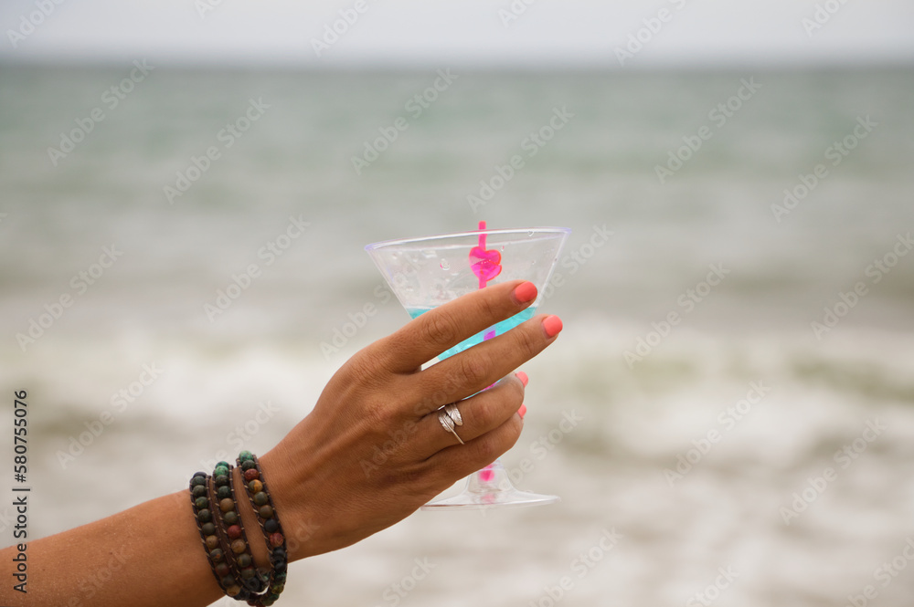 Detail of hand raising glass in the air with a blue drink. In the background you can see the sky and the sea in the horizon.