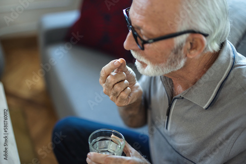 Photo of senior man with glass of water taking pill. Age, medicine, healthcare and people concept. Man taking pill against headaches at home. Gray hair man at home taking pill to ease headache.