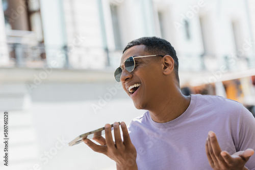 Positive african american man in sunglasses recording voice message on urban street in summer.