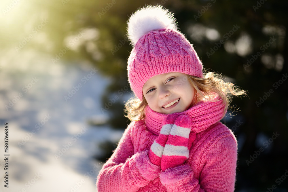 Portrait of a pretty smiling blonde girl with blue eyes, dressed in a knitted hat with a pompom, sweater, scarf and pink mittens. The girl with golden hair folded her ankles and smiles Portrait of a pretty smiling blonde girl with blue eyes, dressed in a knitted hat with a pompom, sweater, scarf and pink mittens. The girl with golden hair folded her ankles and smiles
