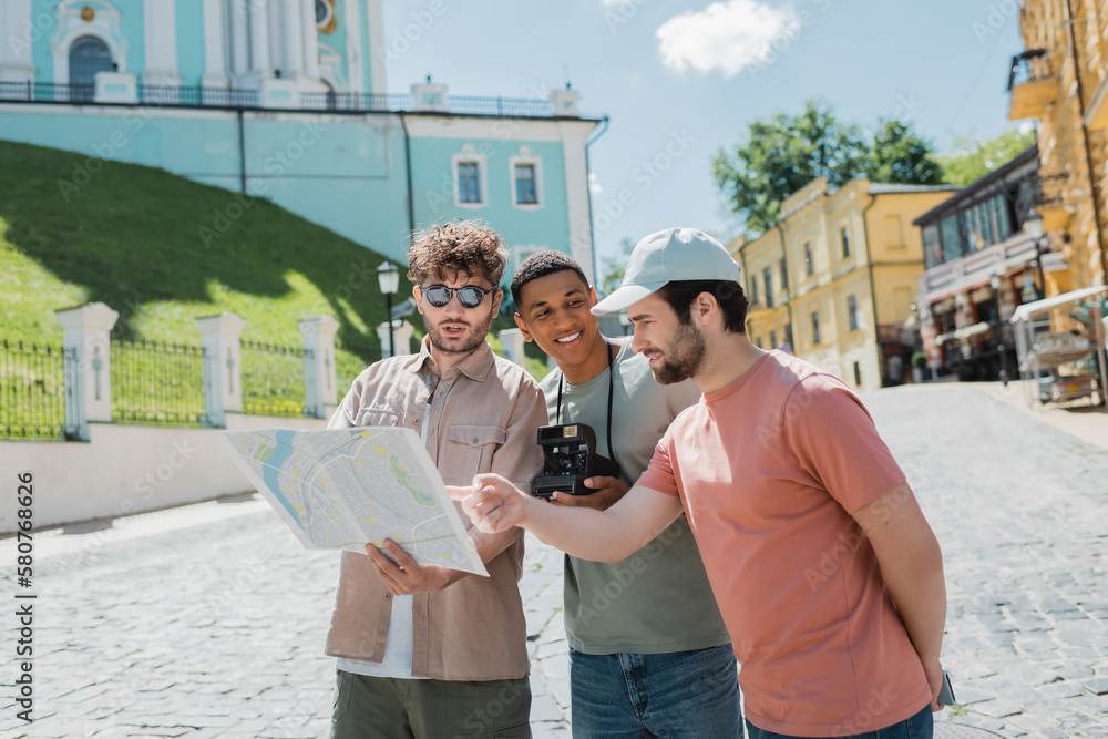 bearded tourist pointing at city map near tour guide and smiling african american man with vintage camera on Andrews descent in Kyiv.