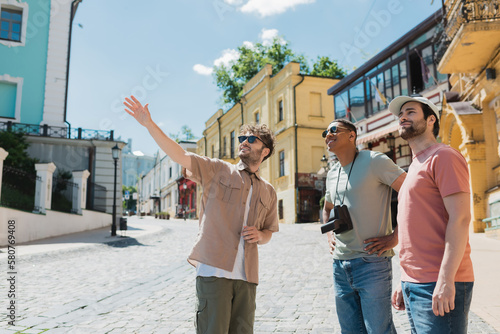 Fotografia tour guide in sunglasses pointing with hand during excursion with interracial tourists on Andrews descent in Kyiv