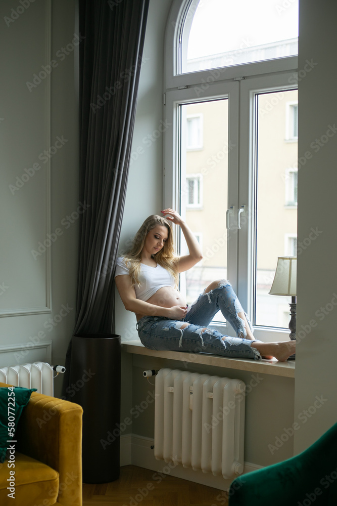 Happy pregnant woman touch bare tummy and look down sitting on windowsill. Home resting, relaxation near window. Happy pregnant woman touch bare tummy and look down sitting on windowsill. Home resting, relaxation near window.