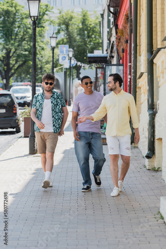 Smiling interracial friends in sunglasses walking on Andrews descent in Kyiv.