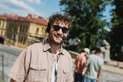 portrait of curly and carefree tour guide in sunglasses and headset smiling near blurred multiethnic travelers on Andrews descent in Kyiv.