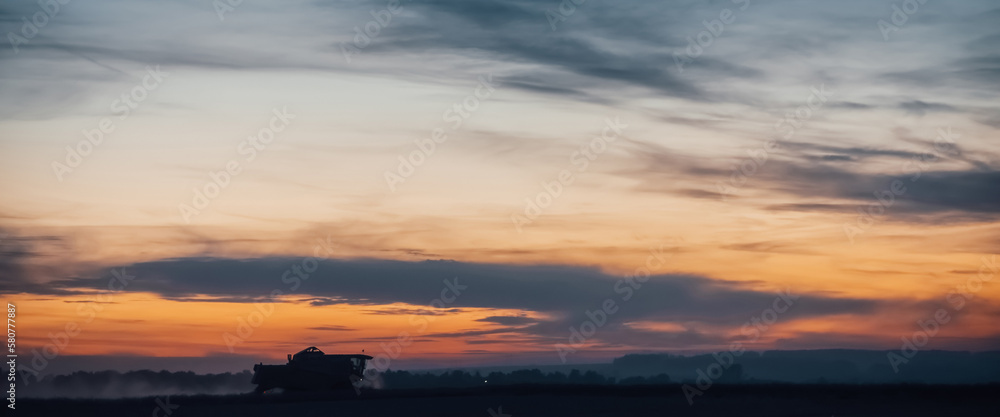 Silhouette of harvester machine to harvest wheat on sunset. Combine harvester driving on field on sunrise. Beautiful dawn sky above wheat field. Combine working in dusk. Wonderful twilight landscape.