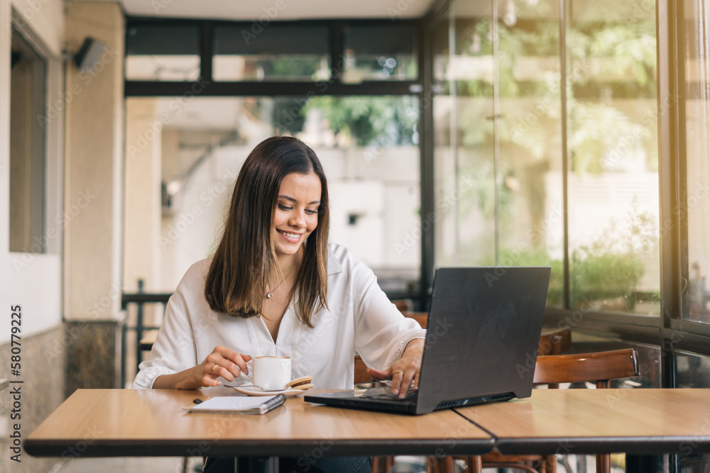 Woman using laptop, communicates on internet with client from coffee shop, reusable coffee/tea mug in hand. Cozy office work, remote work, learning concept Woman using laptop, communicates on internet with client from coffee shop, reusable coffee/tea mug in hand. Cozy office work, remote work, learning concept