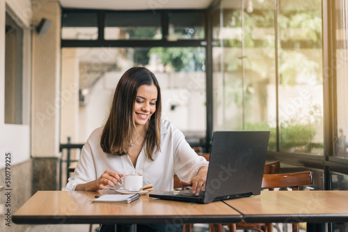Woman using laptop, communicates on internet with client from coffee shop, reusable coffee/tea mug in hand. Cozy office work, remote work, learning concept