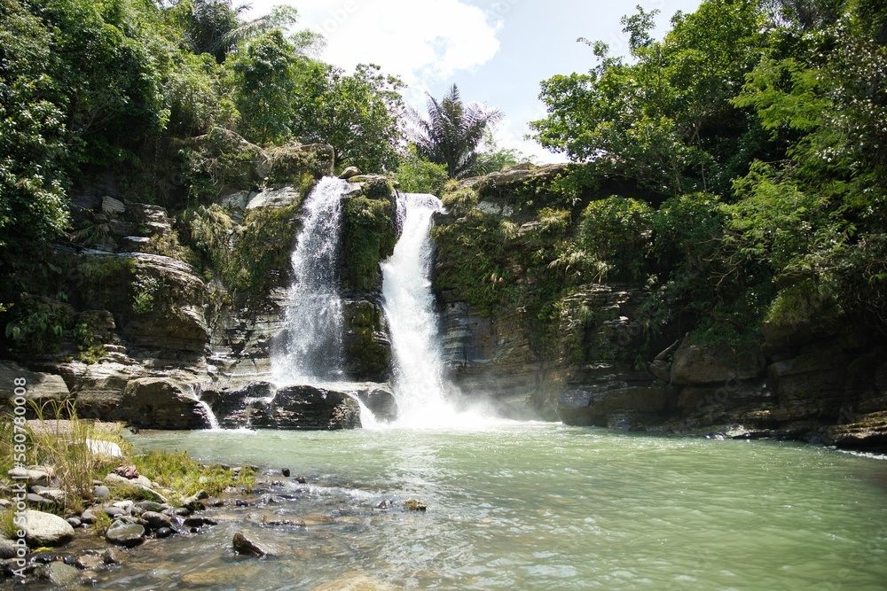 Fototapeta premium The spectacular Air Terjun Tengkulese waterfall on Flores which flows into a pool surrounded by sunlit rainforest.