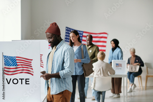 Group of young multicultural voters in casualwear standing in queue along vote booths in polling place and putting their ballots into boxes