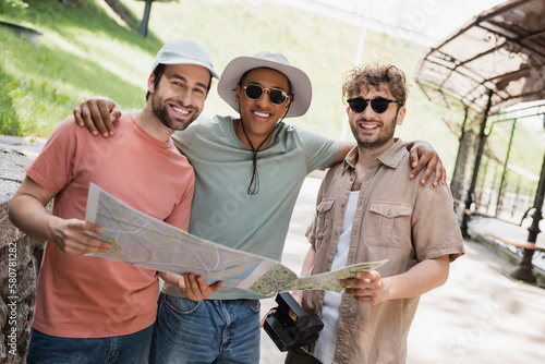 carefree african american man in sun hat and sunglasses embracing friends on urban street.