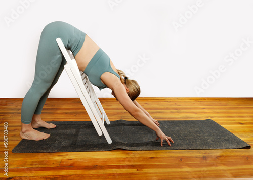 European blonde woman doing yoga in green sportswear practices sports pose indoors with a chair. Young slim woman in sportswear doing yoga for back