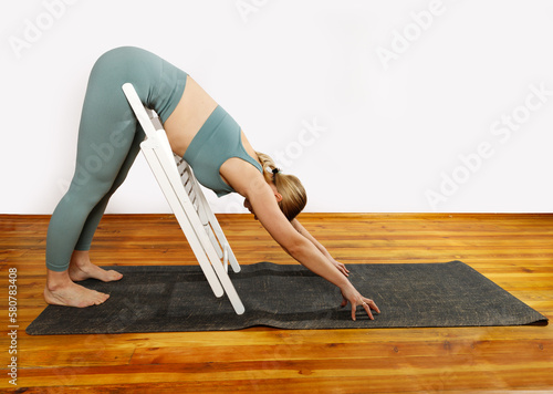European blonde woman doing yoga in green sportswear practices sports pose indoors with a chair. Young slim woman in sportswear doing yoga for back (ID: 580783408)