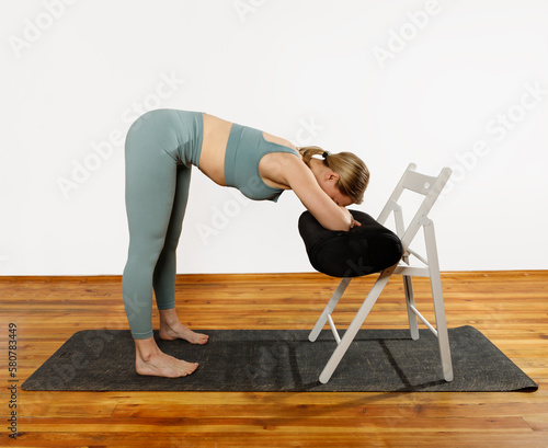 European blonde woman doing yoga in green sportswear practices sports pose indoors with a chair. Young slim woman in sportswear doing yoga for back