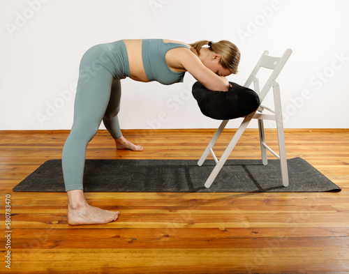 European blonde woman doing yoga in green sportswear practices sports pose indoors with a chair. Young slim woman in sportswear doing yoga for back