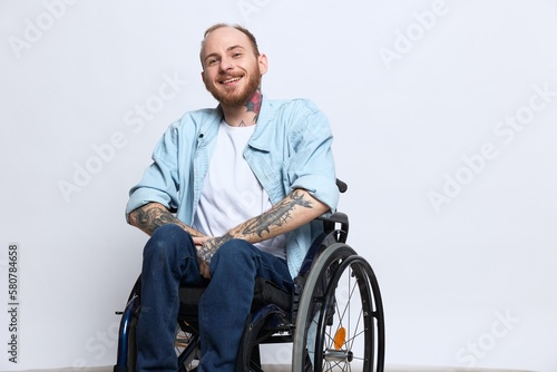 A man in a wheelchair and pointing a finger at, copy space, with tattoos on his hands sits on a gray studio background, health concept a person with disabilities, a real person