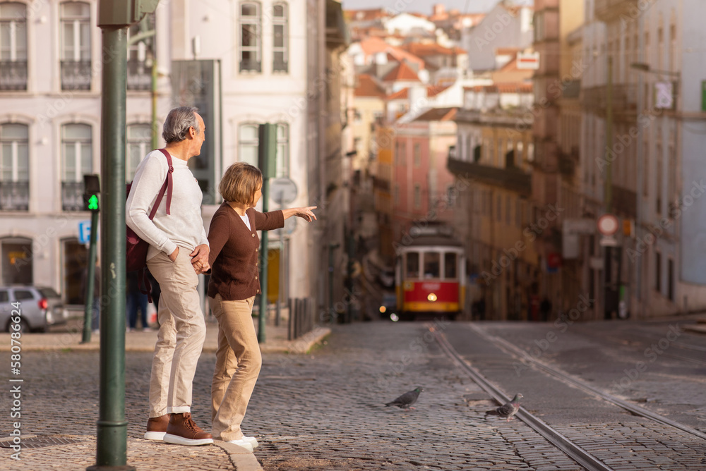 Senior Tourists Couple Walking In Lisbon, Pointing Finger At Tram