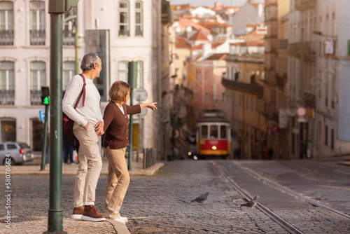 Senior Tourists Couple Walking In Lisbon, Pointing Finger At Tram
