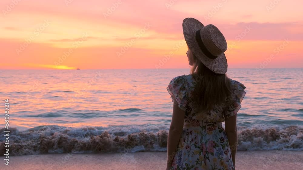 A woman in a flowing dress walks on a paradise beach at golden pink ...