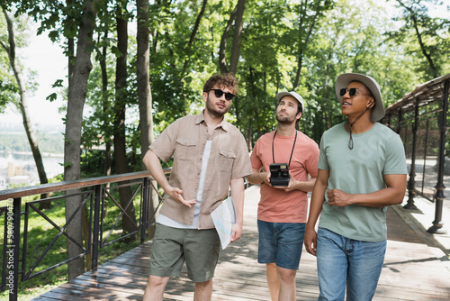 multicultural travelers in sun hats looking away during summer walk with tour guide in urban park.