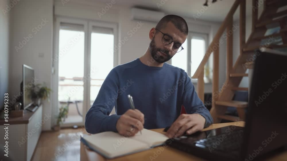 Professional focused and happy man working from home. Employee sit at desk at home working with laptop and papers, writes down ideas make notes and using phone Professional focused and happy man working from home. Employee sit at desk at home working with laptop and papers, writes down ideas make notes and using phone