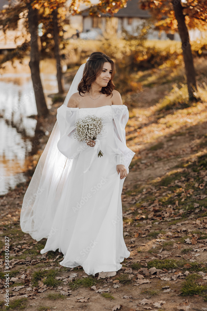 Awesome bride in a long wedding dress with a beautiful bouquet in hand and with charming smile. Under the tree in the beautiful sunset light in the park.