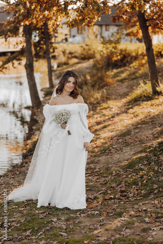 Awesome bride in a long wedding dress with a beautiful bouquet in hand and with charming smile. Under the tree in the beautiful sunset light in the park.