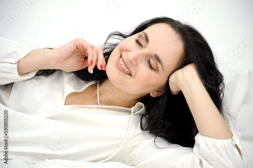 pleasure bliss pleasant emotions beautiful brunette woman lies on white bed in pajamas in nightgown throwing head back closed her eyes smiling tenderness self-care facial relaxation rest hotel