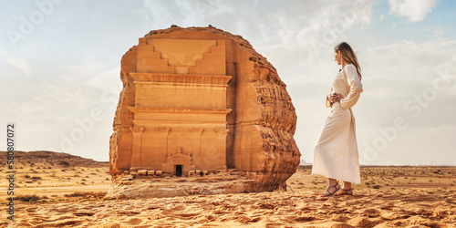 Young Caucasian woman in long white dress posing in front Tomb Lihyan Son of Kuza or Qasr al-Farid at Hegra, Saudia Arabia, sandy desert landscape around. Saudi Arabia opened to tourism in 2019 photo