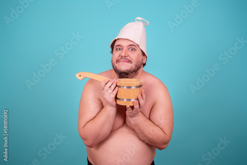 Funny fat man in a hat and with a sauna ladle. Blue background. Taking a shower.