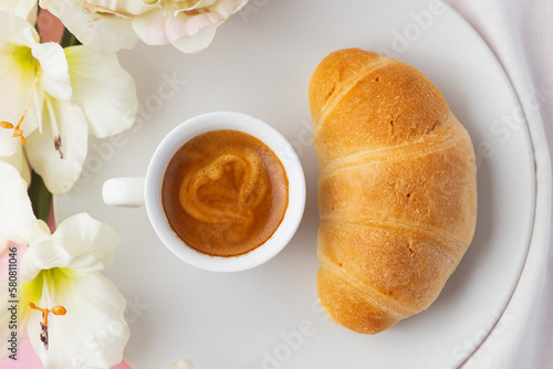 Top close upview of croissant on a white plate with a cup of espresso coffee with a heart on it, flowers and pink background photo