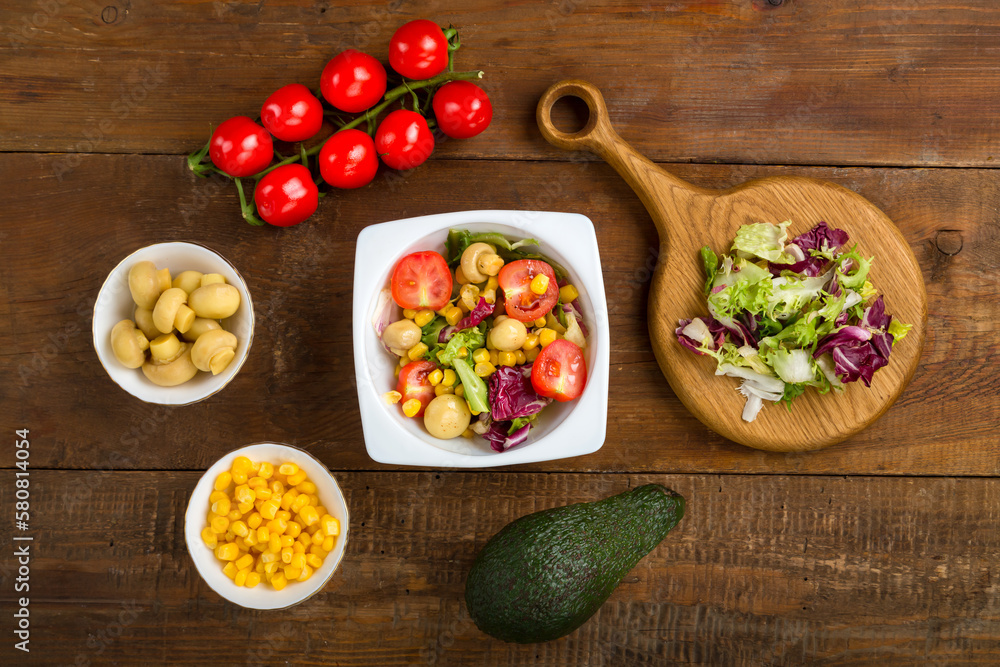 Vegetable salad with champignon corn and cherry tomato with avocado in a white plate on a wooden table next to all the ingredients. Vegetable salad with champignon corn and cherry tomato with avocado in a white plate on a wooden table next to all the ingredients.
