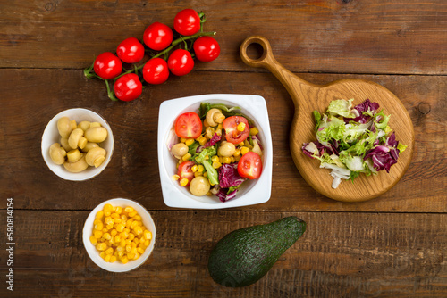 Vegetable salad with champignon corn and cherry tomato with avocado in a white plate on a wooden table next to all the ingredients.