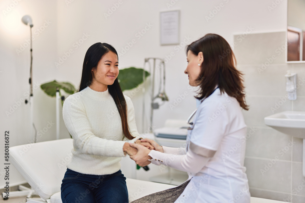 A smiling Asian female patient holding hands with a female doctor at the clinic. A smiling Asian female patient holding hands with a female doctor at the clinic.