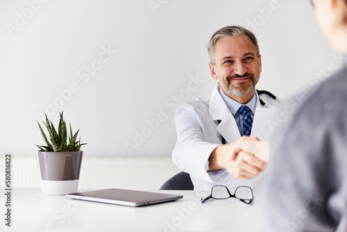 A senior doctor in a white coat, sitting at the office and handshake with a patient.