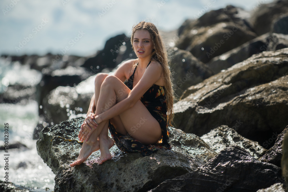 Sexy woman sit on stone rocks on beach. Traveller woman summer dress on beautiful sandy beach. Cute girl enjoy her tropical sea on relax holiday vacation during summer day.