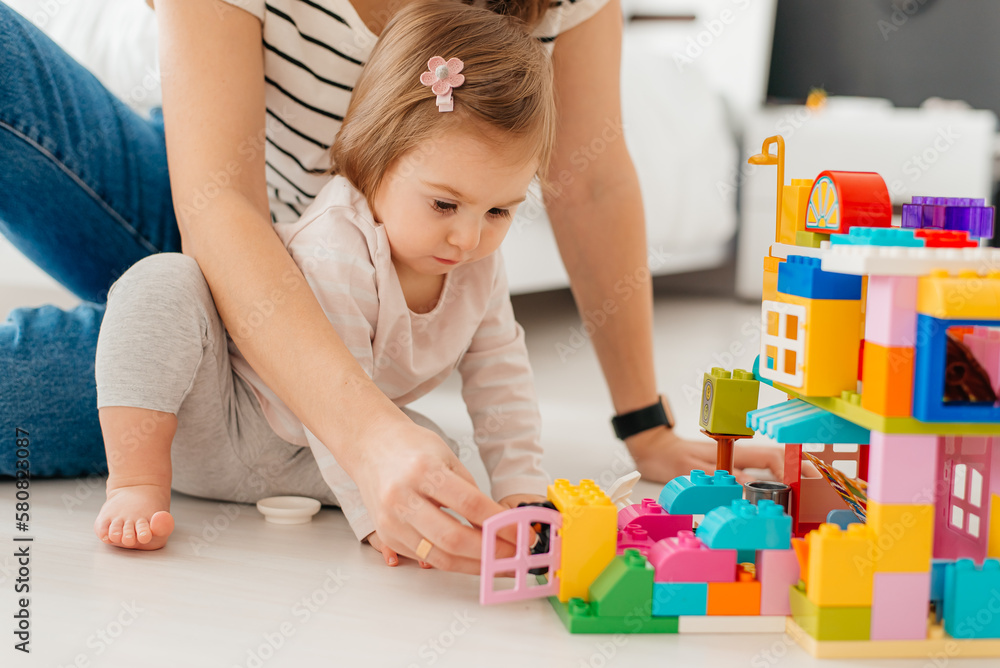 Laughing mother and little daughter playing colorful blocks, constructing tower, sitting on warm floor with underfloor heating, family enjoying leisure time together Laughing mother and little daughter playing colorful blocks, constructing tower, sitting on warm floor with underfloor heating, family enjoying leisure time together