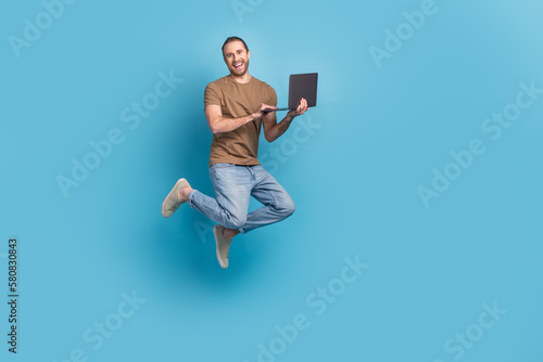 Full length photo of cute funny man wear beige t-shirt jumping high communicating modern device isolated blue color background