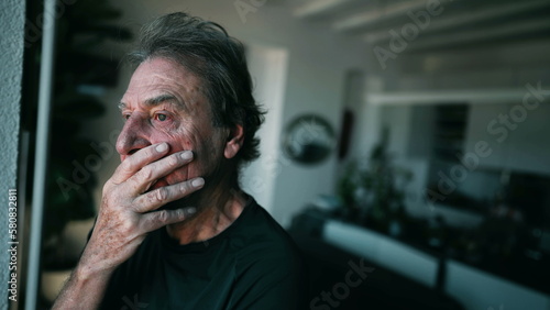 One depressed senior man thinking about problems at home living room. Mature male person in 70s with desperate emotion stands by window indoors