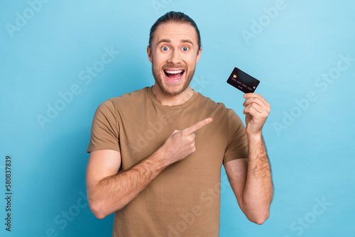 Photo of entrepreneur man wear t-shirt direct finger surprised demonstrate plastic credit card wireless nfc payment isolated on blue color background